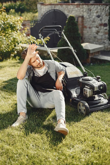 A young man sitting on a well-maintained grassy lawn next to a petrol-powered lawn mower, with his hand resting on his forehead in a gesture of fatigue or frustration. The garden features lush green grass, surrounding garden beds with flowering shrubs, and a stone pathway leading to a dark wooden gate set into a brick wall in the background. There are trimmed hedges and tall trees, providing shade and a scenic backdrop under clear sunlight. The scene suggests a setting of outdoor gardening or lawn care, with the man appearing to take a break after mowing or edging the lawn. The environment is neatly landscaped, highlighting tools and freshly cut grass typical of regular garden maintenance, and the overall atmosphere emphasizes attention to landscape upkeep, consistent with the services offered by Gardeners Sutton.