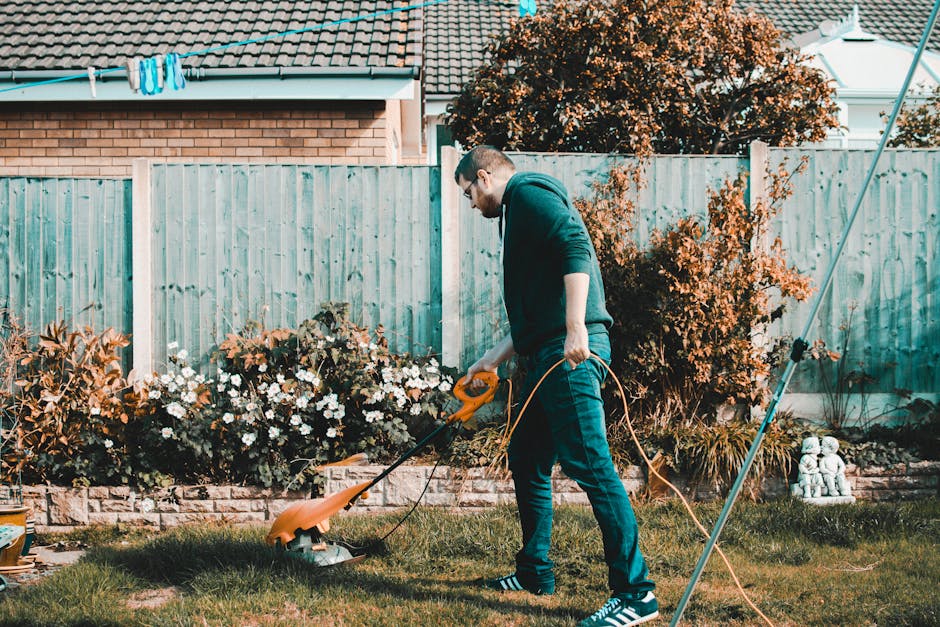 A man wearing glasses, a dark hoodie, and jeans is trimming the grass with a yellow electric lawn mower in a well-maintained garden. The garden features a lush green lawn, bordered by flower beds filled with white and purple flowers, and edged with a low brick wall. Behind him, there are neatly trimmed shrubs and a tall hedge alongside a light blue wooden fence, with a row of trees and a house with a tiled roof in the background. The scene is lit by natural daylight, highlighting the textures of the wet soil, trimmed grass, and healthy plants, indicating ongoing garden maintenance work. Gardeners Sutton’s presence is implied through the context of professional outdoor care, focused on lawn care and hedge trimming, in a tidy, landscaped outdoor space.